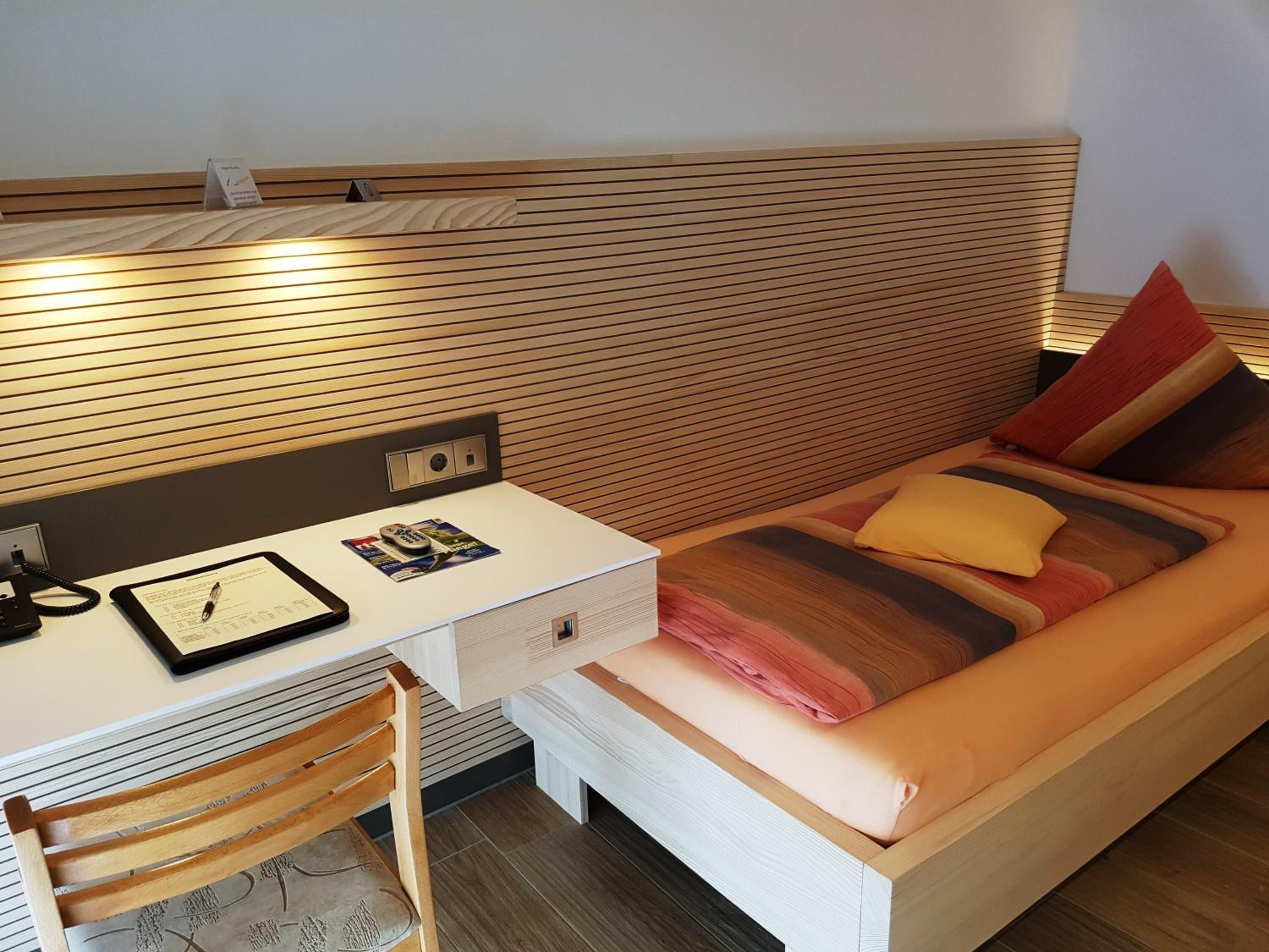 A compact hotel room with a wooden headboard, a desk with a telephone and a tablet, a bed with striped bedding, and a wooden chair.