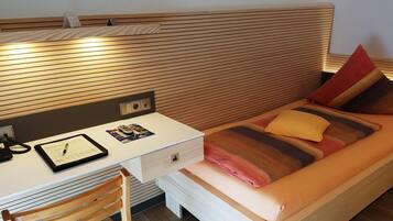 A compact hotel room with a wooden headboard, a desk with a telephone and a tablet, a bed with striped bedding, and a wooden chair.