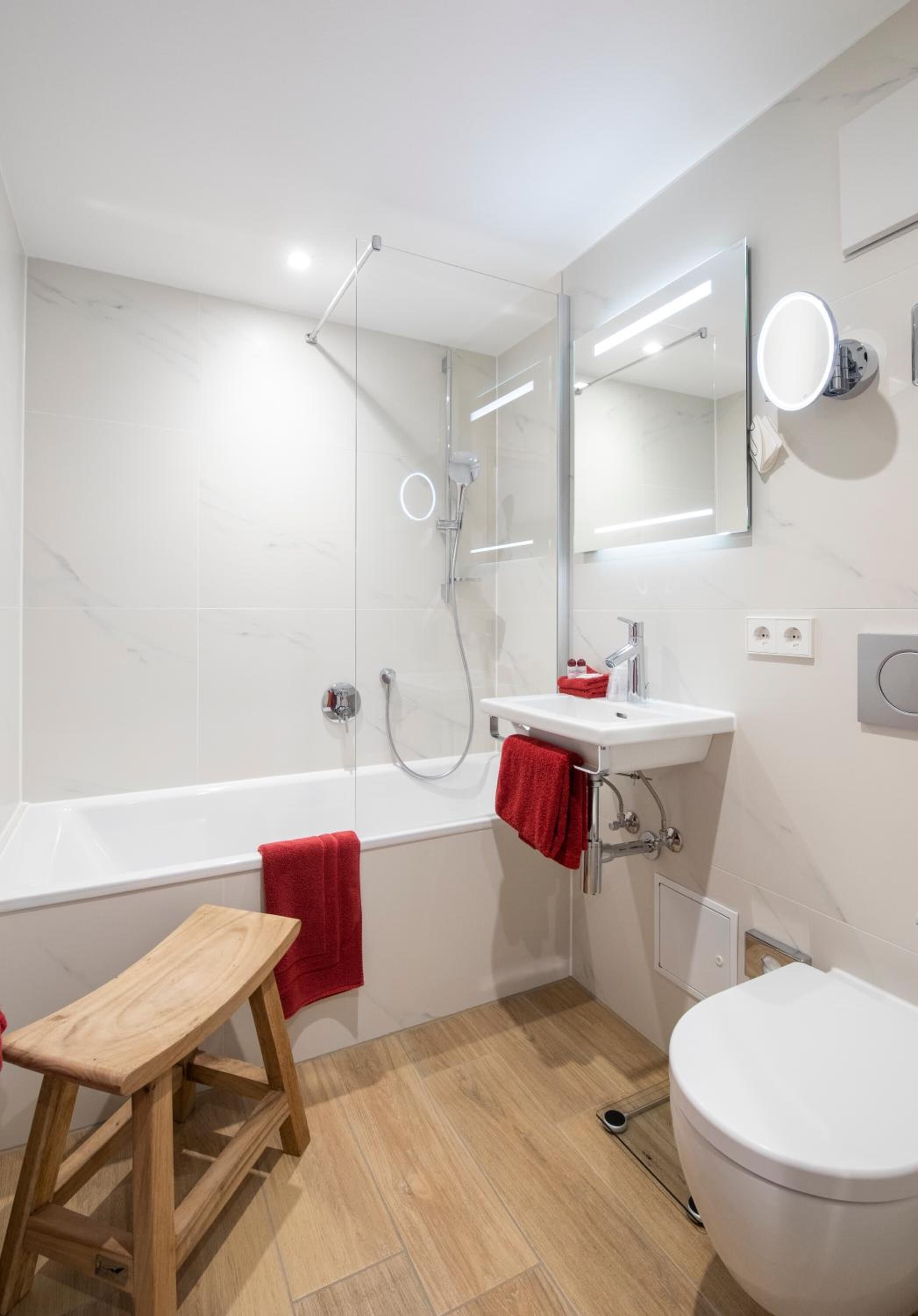A modern bathroom with a glass-enclosed shower, a white sink with a mirror, a wooden stool, and a toilet.