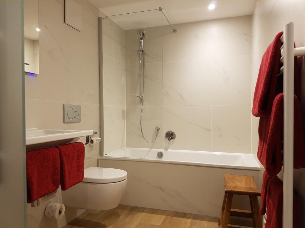 A modern bathroom with a toilet, a shower, and a bathtub. There are red towels hanging on the wall and a wooden stool.