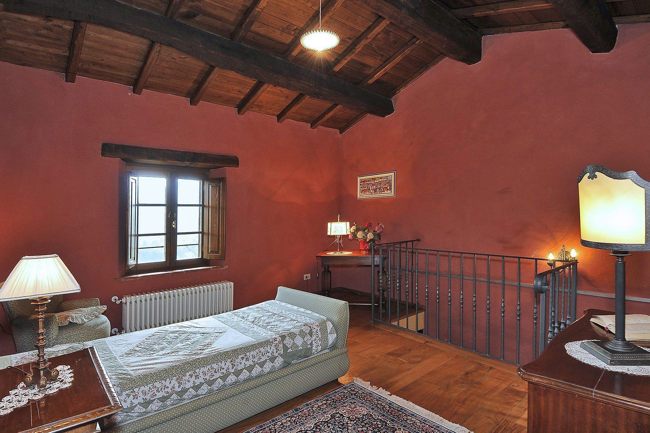 Une chambre avec un plafond en bois, un lit recouvert d’une couvre-lit à motifs, une table de chevet avec une lampe, une fenêtre et un radiateur.