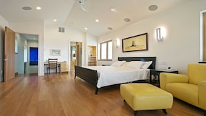A spacious bedroom with a large bed, a desk, and a chair, a yellow ottoman, and a painting on the wall.
