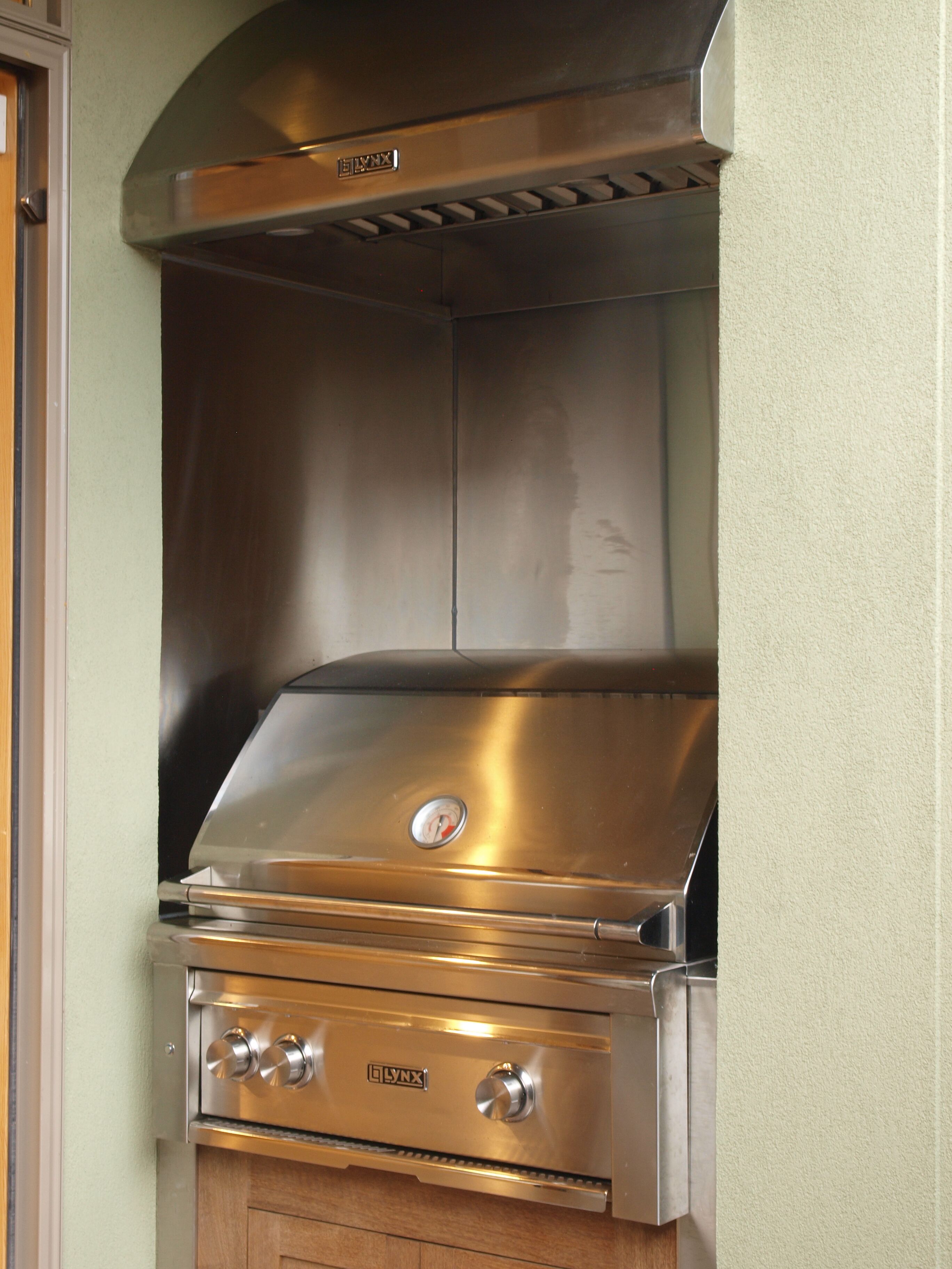A stainless steel LYNX gas grill with a hood and control knobs, installed in a kitchen with wooden cabinets.