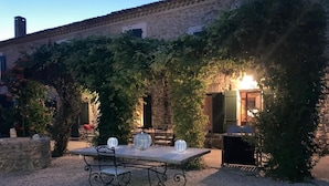 An outdoor dining area with a table and chairs, surrounded by greenery and a stone building in the background. - Mas La Pomarède (Robion)
