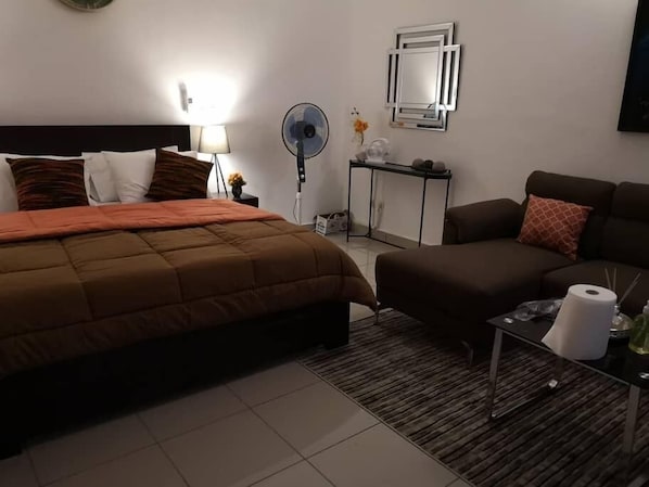 A hotel room with a bed, a sofa, a nightstand, a lamp, a fan, a mirror, and a small table with a bottle.