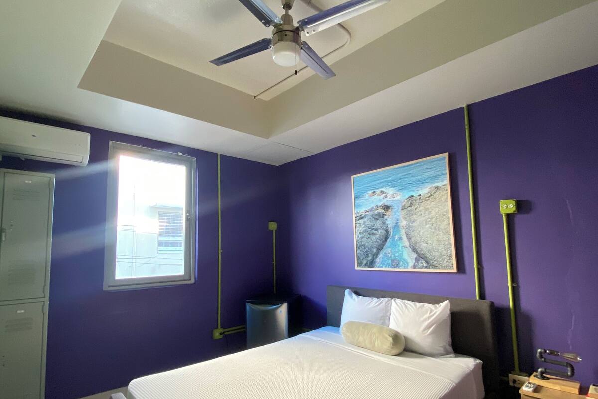 Un dormitorio con cama, ventilador de techo, ventana y un cuadro en la pared.