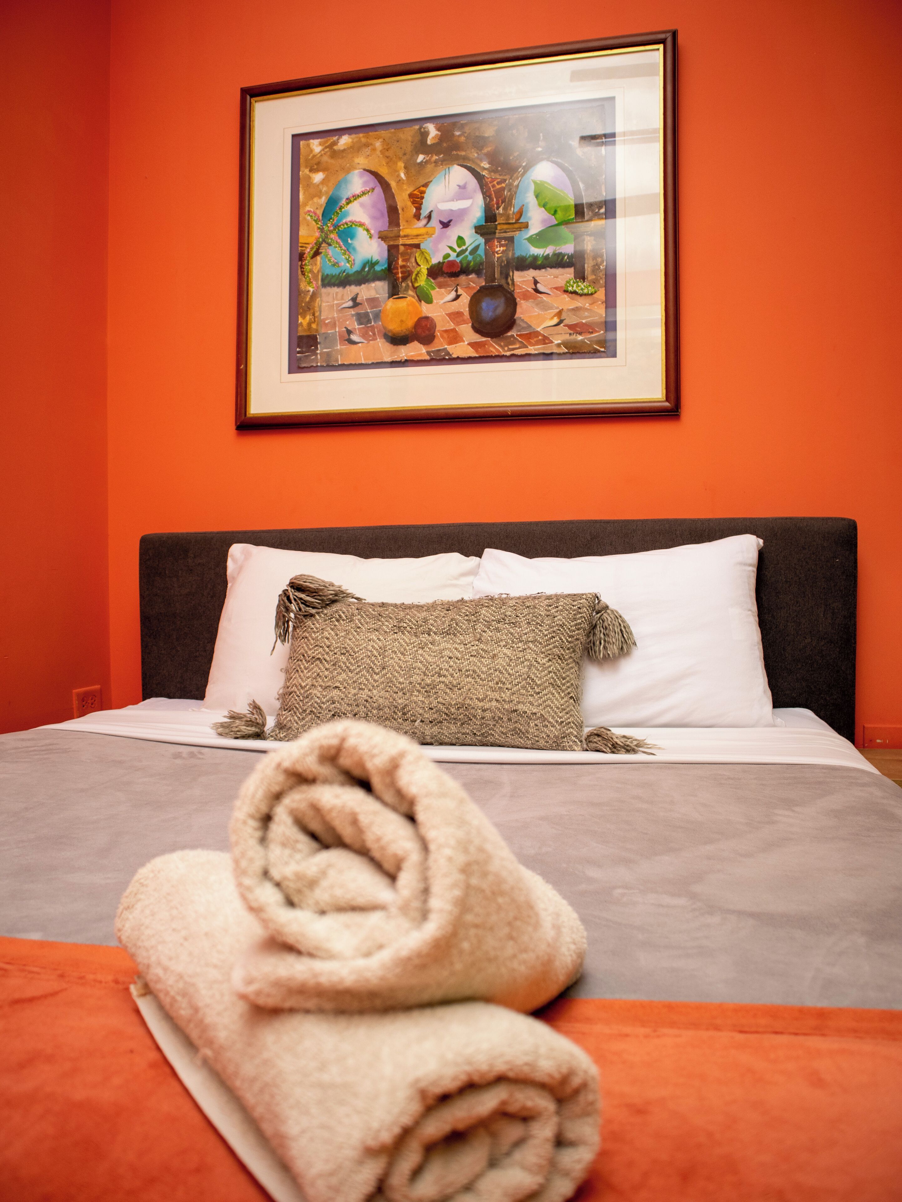 Ein ordentlich gemachtes Bett mit einem zusammengerollten Handtuch, ein gerahmtes Bild darüber und einer schlichten orangefarbenen Wand.
