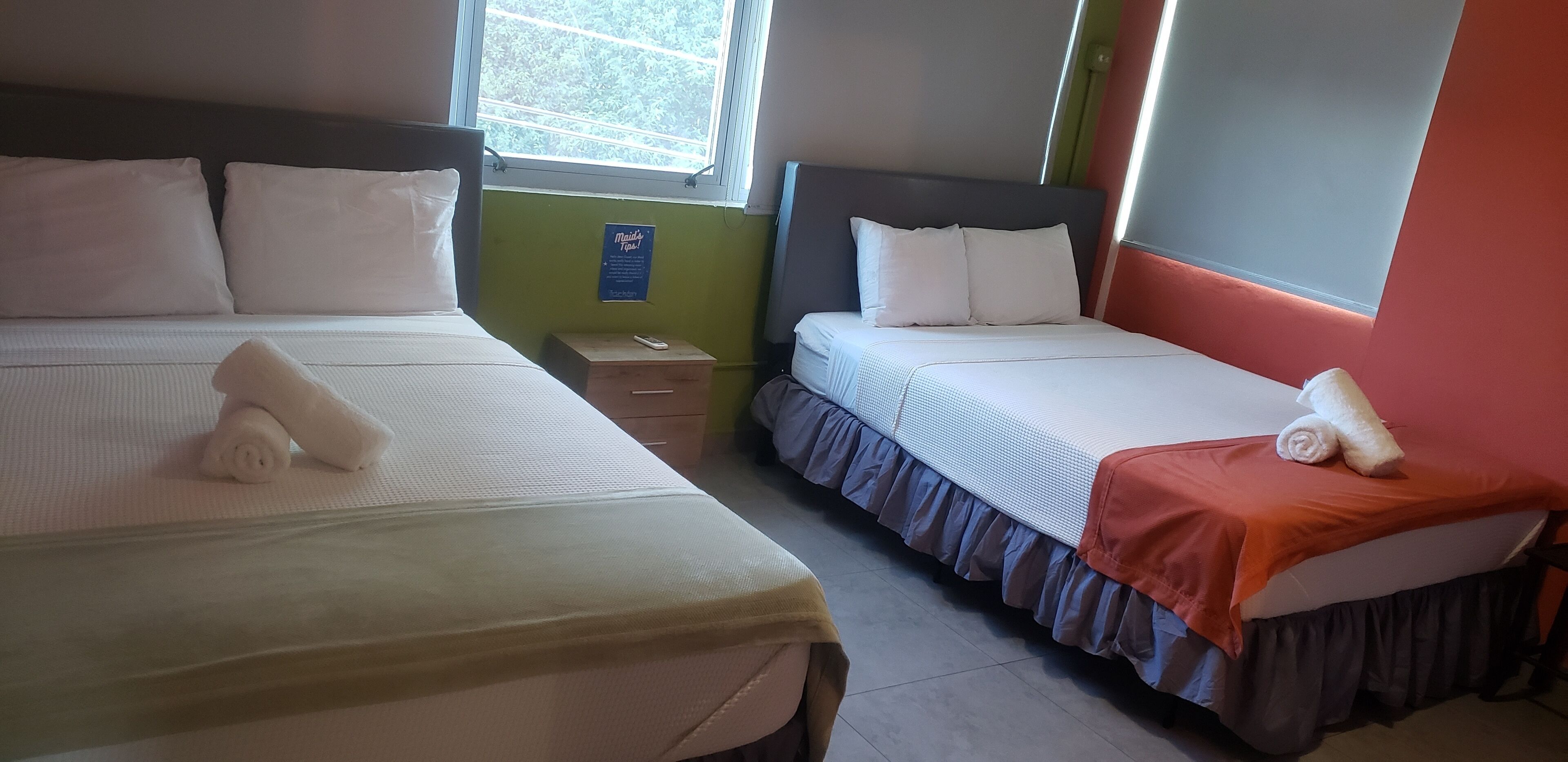 Habitación de hotel con dos camas, cada una con sábanas y almohadas blancas.  Una pequeña mesa entre las camas sostiene un control remoto.