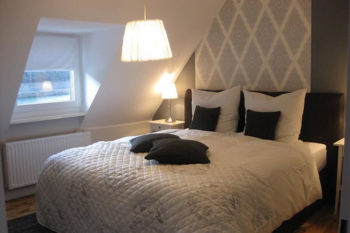 A bedroom with a large bed, a nightstand, a lamp, a window, and a patterned wall.