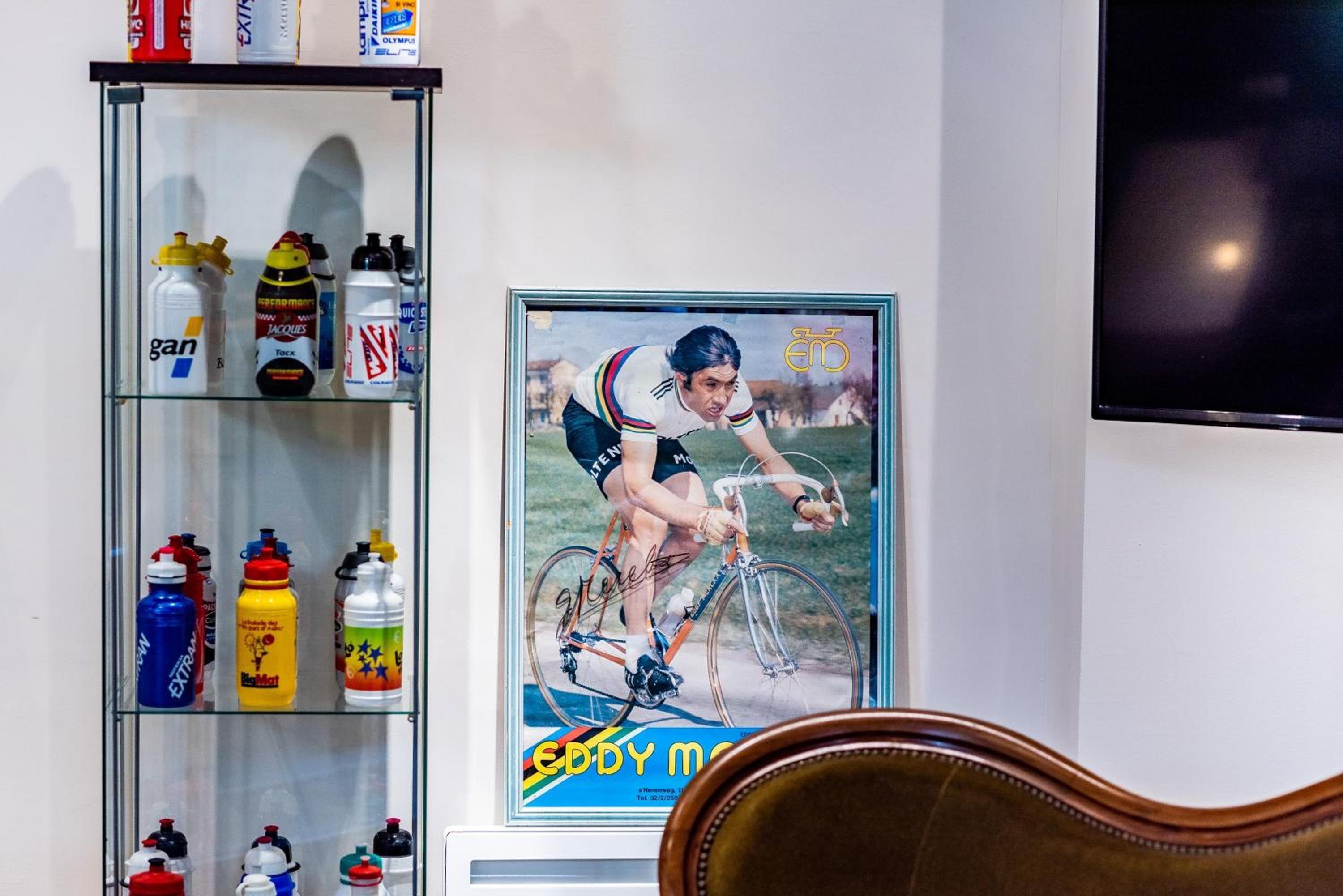 Una habitación con un mueble de vidrio que contiene varias botellas de agua, un cartel de ciclismo enmarcado y un televisor montado en la pared.