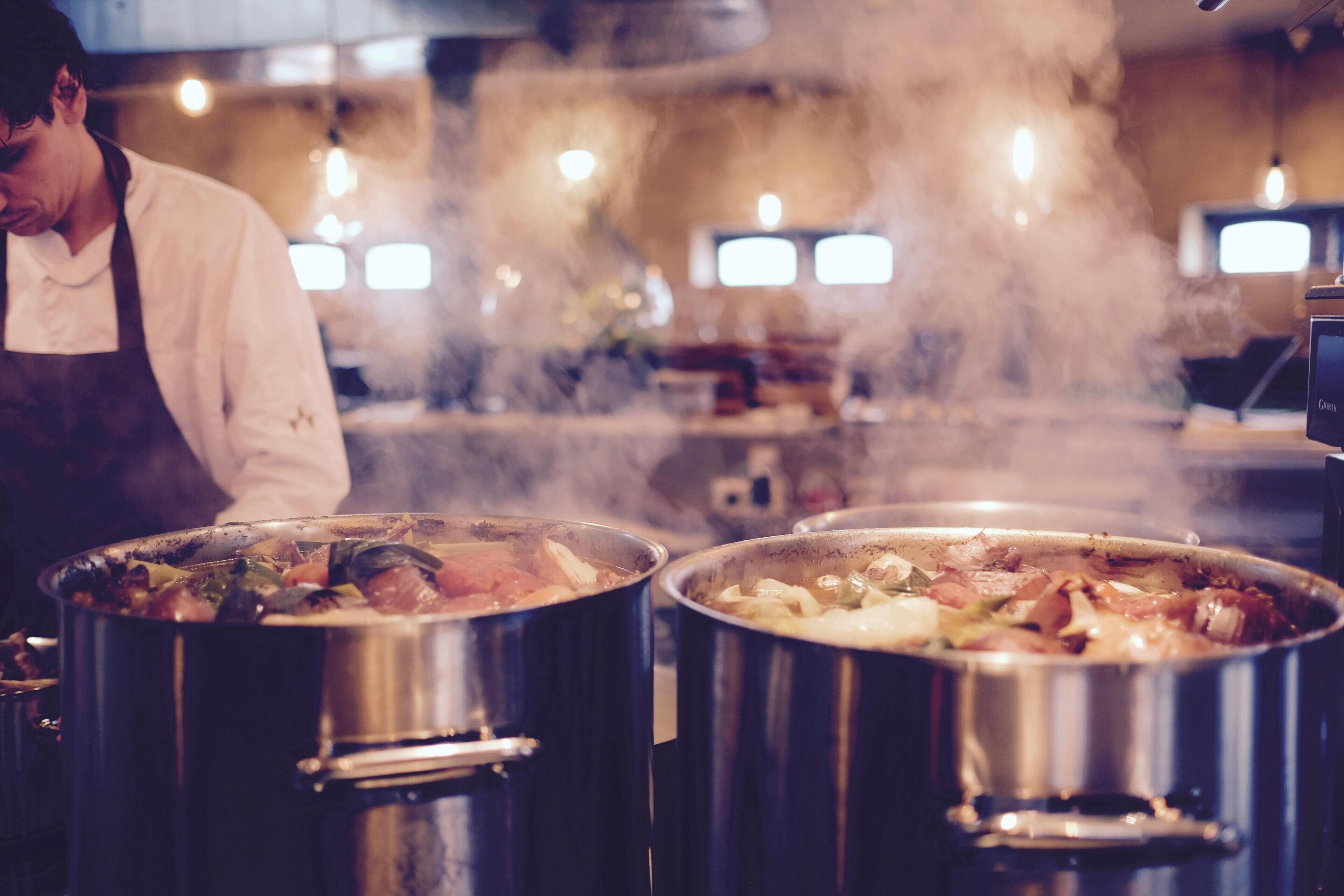 A chef in a kitchen with two large pots on the stove, steam rising, and a television in the background.