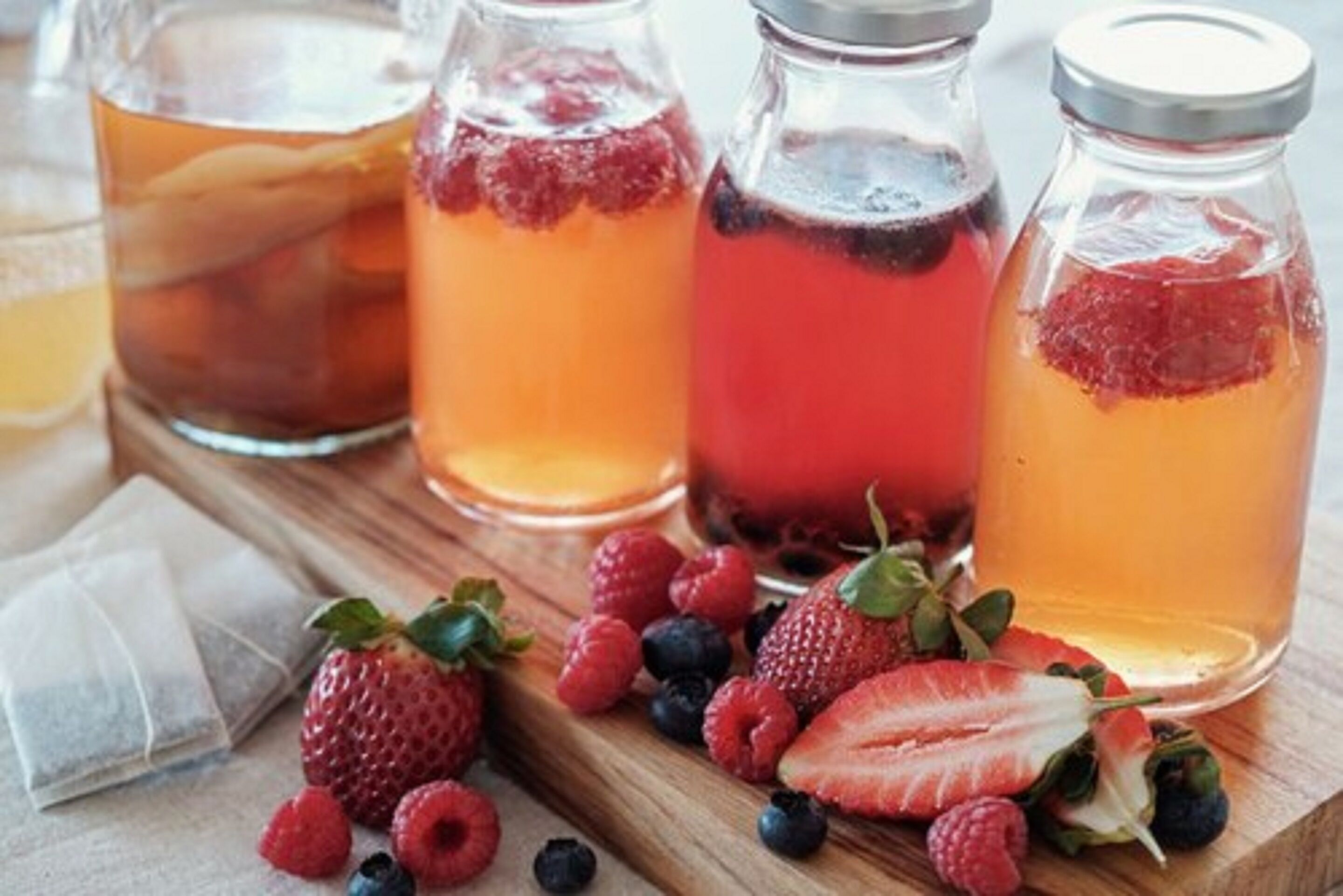 Four glass bottles with different colored liquids, possibly fruit juices, and fresh berries like strawberries and raspberries on a wooden surface.