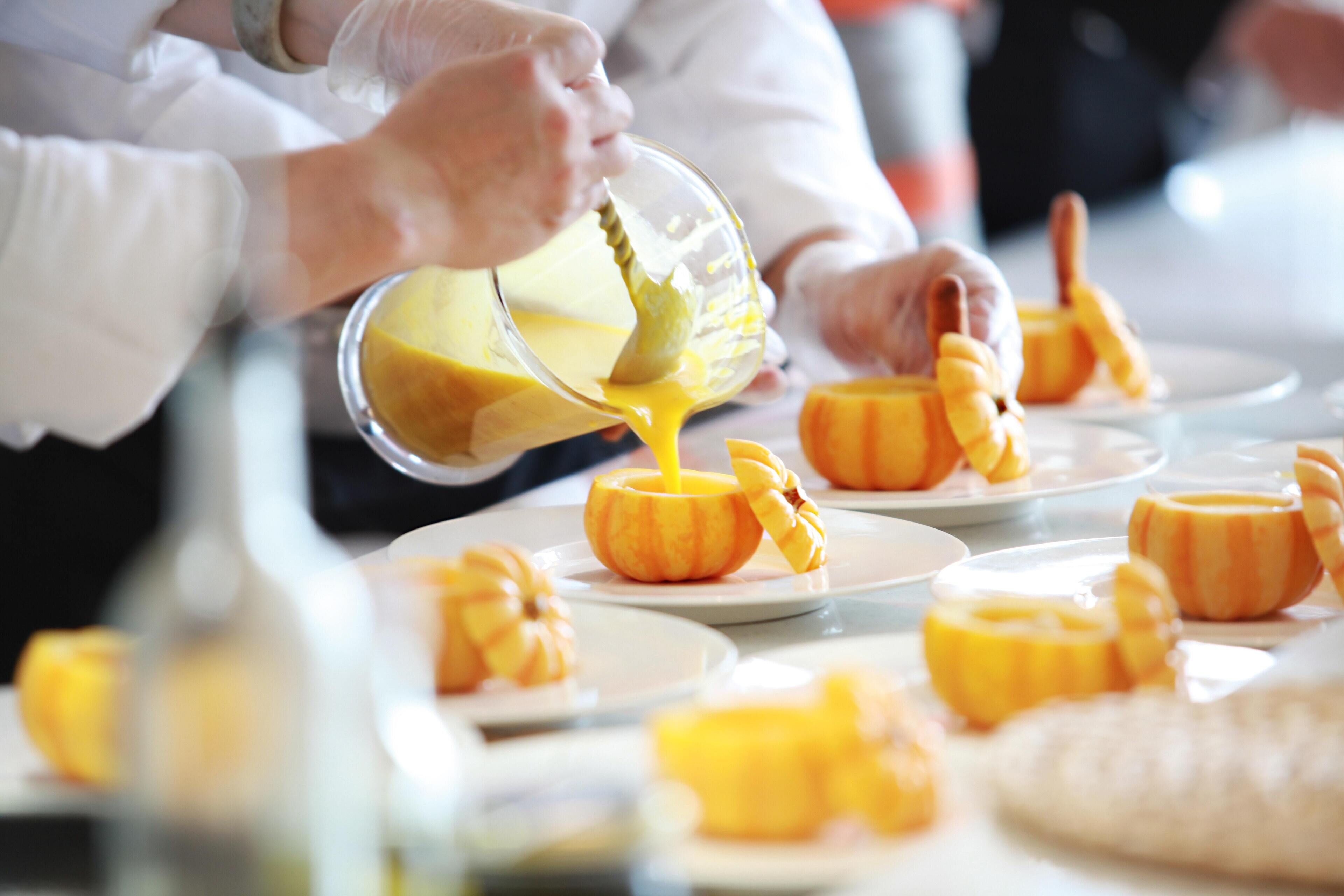 A person pouring a yellow liquid into carved pumpkin bowls on a table.