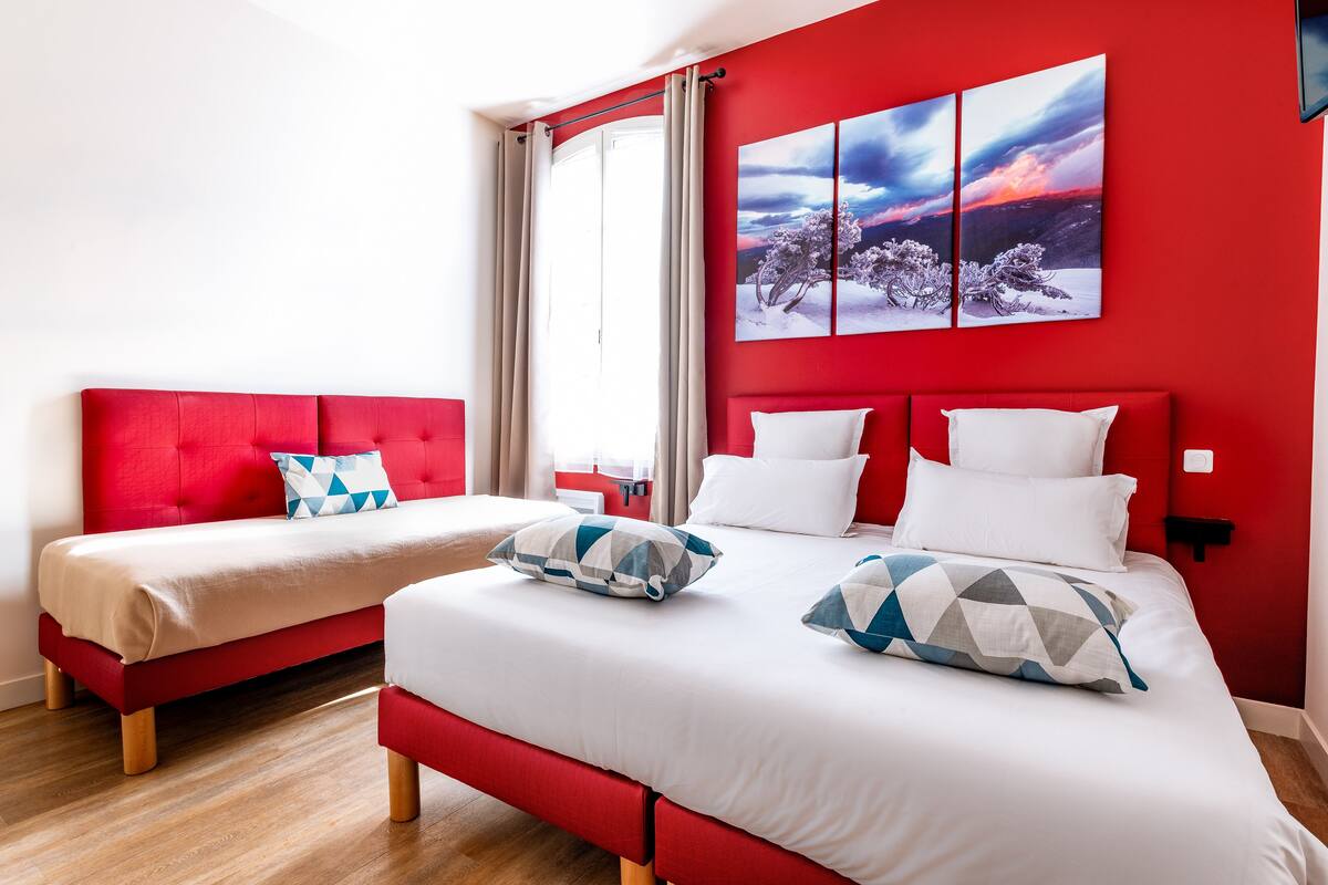 Une chambre d’hôtel avec deux lits, une literie rouge et blanche, et une grande fenêtre avec des rideaux.