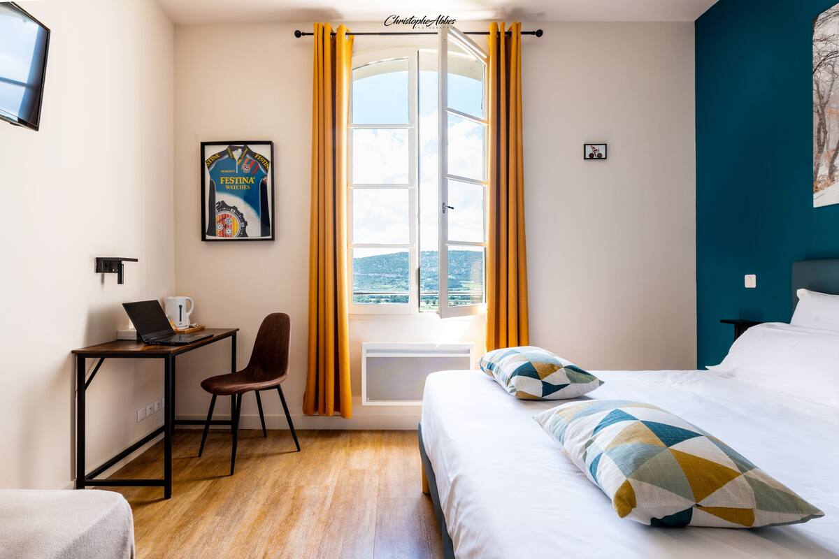 Une chambre d’hôtel avec un lit, un bureau avec un ordinateur portable, une chaise, une fenêtre avec des rideaux et une affiche au mur.