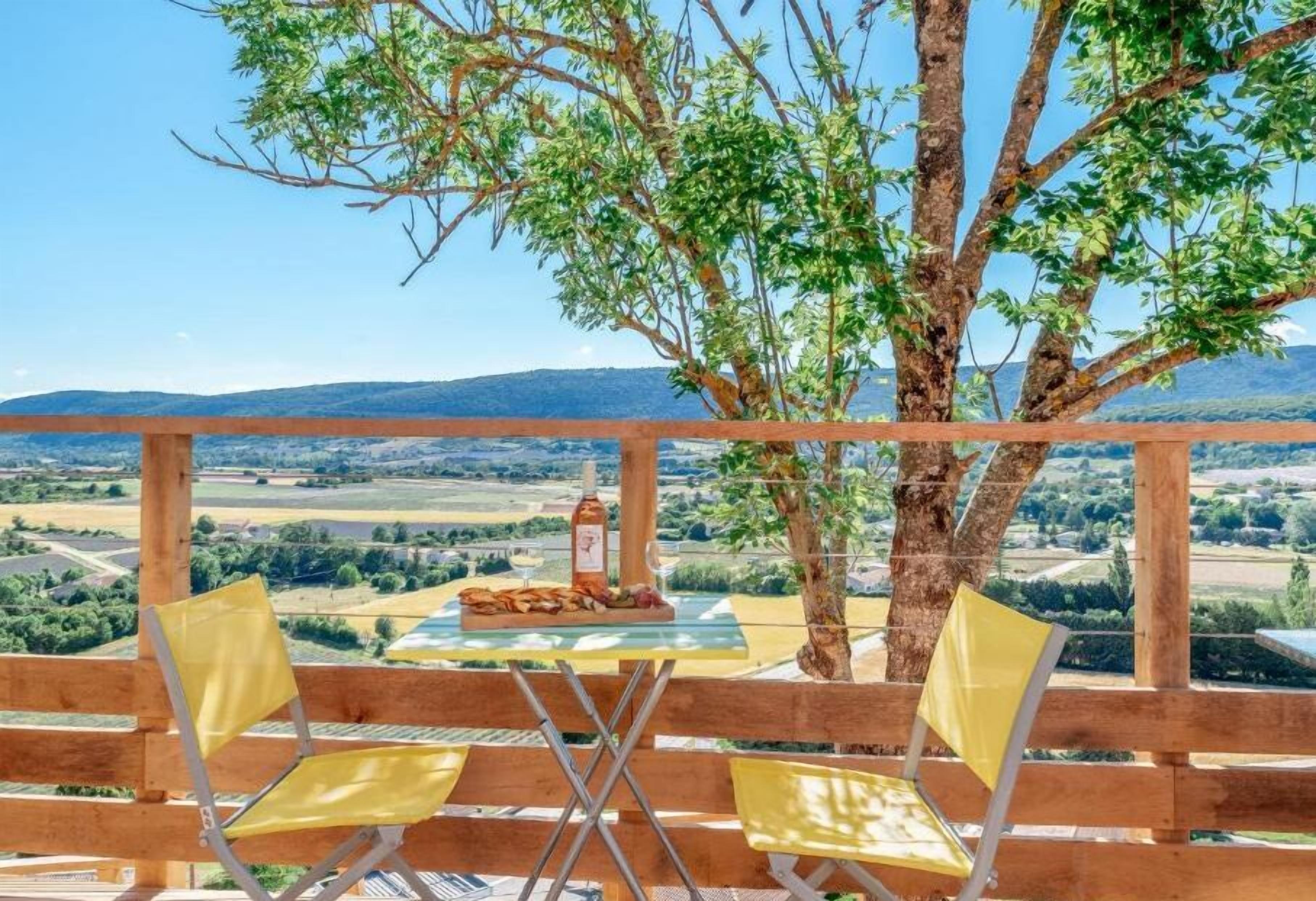 A wooden deck with two yellow chairs and a table set for a meal, overlooking a scenic landscape.