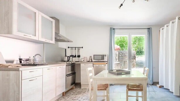 A modern kitchen with white cabinets, a dining table, and a view of the outdoors through a glass door. - The Maison Rose studio (Callas)