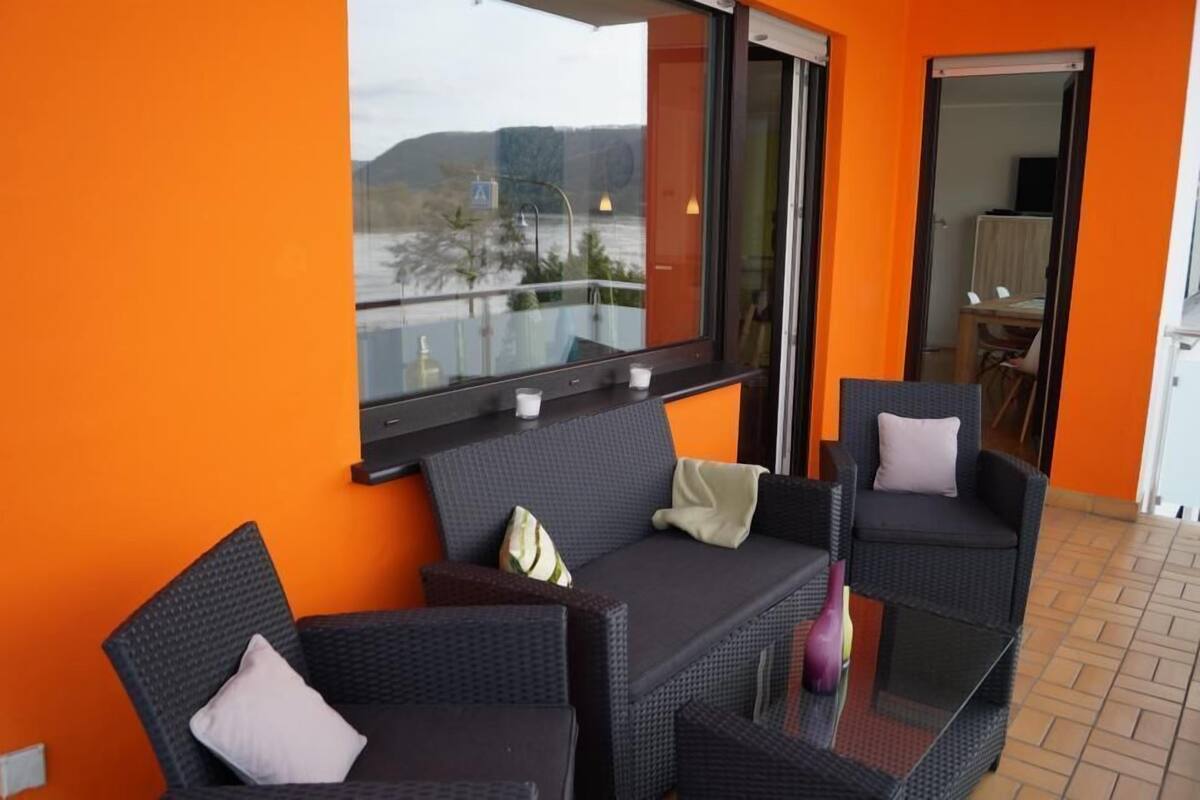 A balcony with wicker furniture, a glass table, and a view of a body of water and hills.