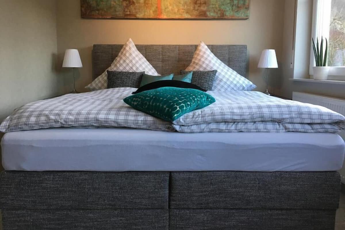 Un lit bien fait, avec une couette à carreaux, plusieurs oreillers et un coussin vert qui apporte une touche de couleur.