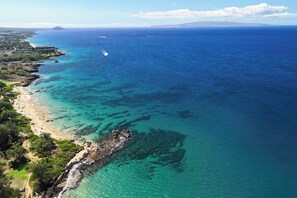 An aerial view of a coastal area with a beach, clear blue waters, and a rocky shoreline.