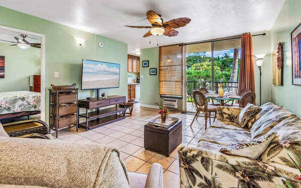 A living room with a sofa, a TV, a dining table with chairs, a ceiling fan, and a view of the outdoors through a sliding door.