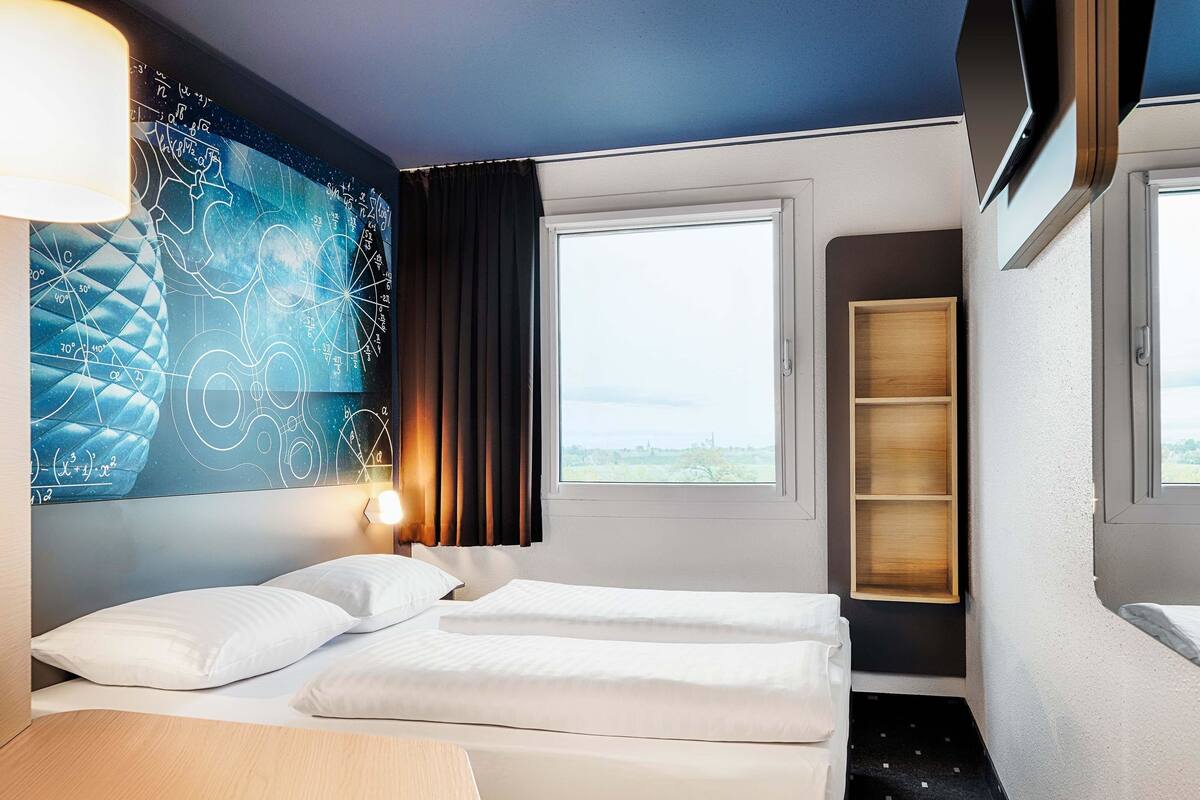 A hotel room with two beds, a large window, a wall with a geometric design, and a built-in shelf.