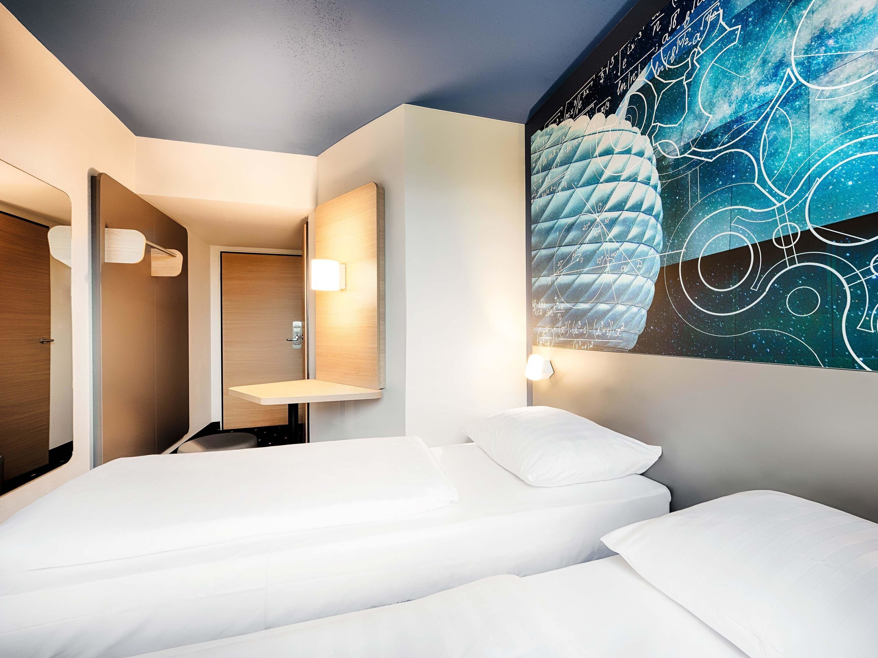A hotel room with two beds, a wooden door, a mirror, and a wall with a geometric design.