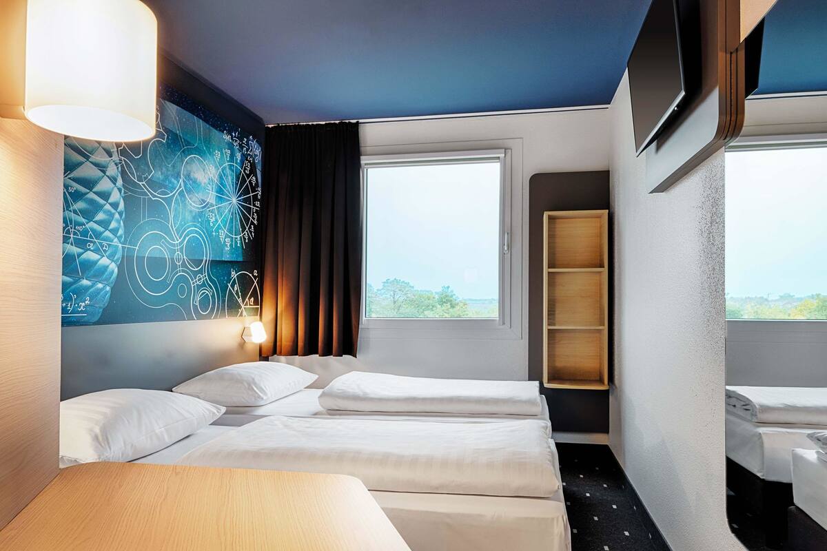 A hotel room with two beds, a large window, a wall tapestry, and a wooden shelf.