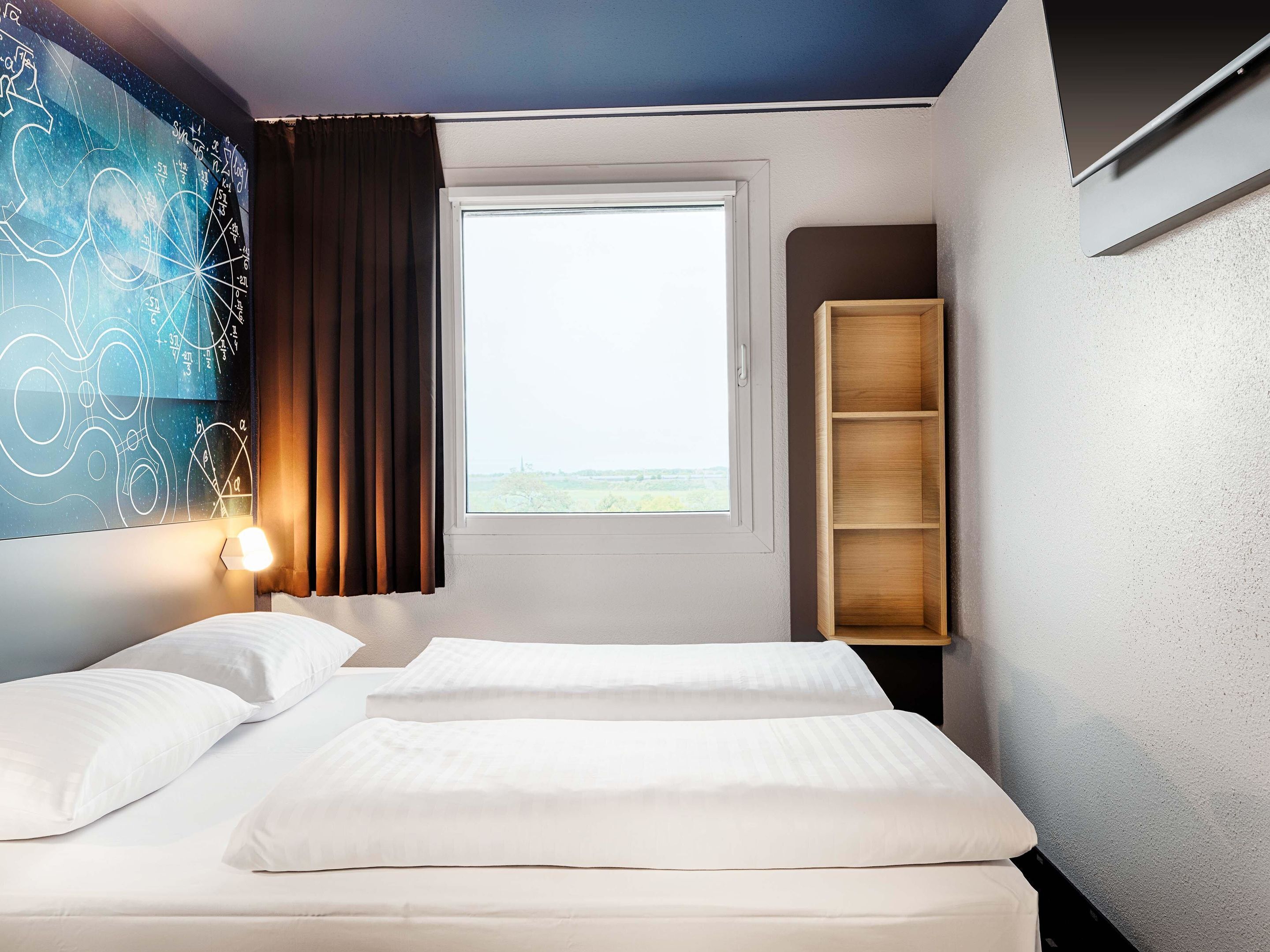 A hotel room with two beds, a window with a view, a wall with a geometric design, and a wooden shelf.