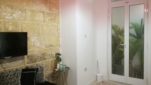 Una habitación con pared de ladrillo, un televisor de pantalla plana montado en ella, un sofá negro con cojines estampados, una mesita de cristal con una planta, y un ventanal corredero que da a un balcón con plantas.