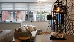 Una sala de estar moderna con sofá, mesa de centro, mesa de comedor y sillas. Hay una ventana con persianas, una lámpara de techo y un estante en la pared con objetos decorativos.