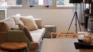 Una sala de estar moderna con un sofá, una mesa de comedor y sillas. Hay una ventana con persianas, una lámpara de pie y una estantería.