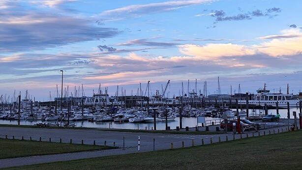 A marina with numerous boats docked, a large building in the background, and a clear sky with clouds. - romantic Oberstübchen (Norden)