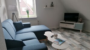 A modern living room with a sectional sofa, a small white coffee table, a television on a white cabinet, and a window with a view of a building. - romantic Oberstübchen (Norden)