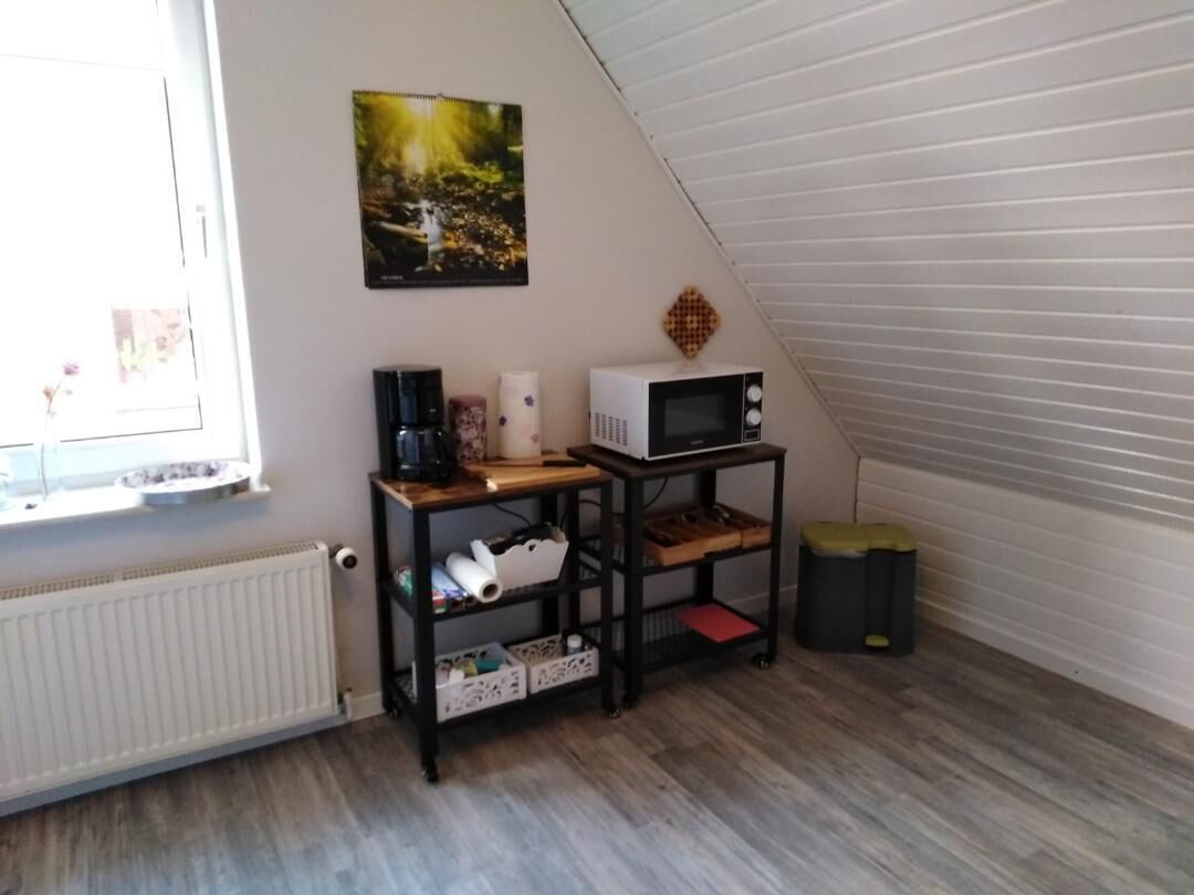 A small, cozy room with a sloped ceiling, a radiator, a wooden shelf unit, a microwave, and a trash bin.