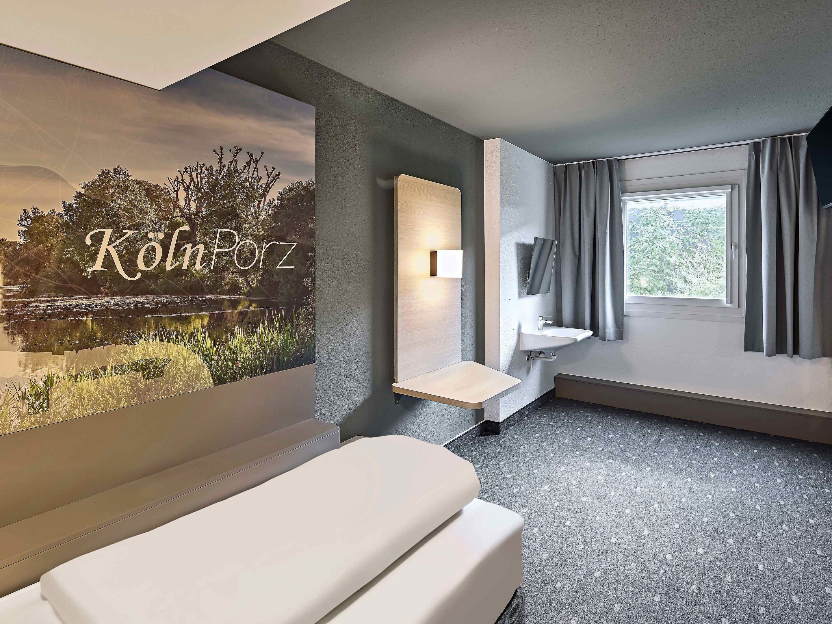 A modern hotel room with a large window, a bedside table, a wall-mounted mirror, and a poster of a lake.