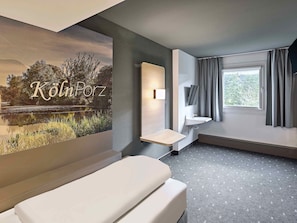 A modern hotel room with a large window, a bedside table, a wall-mounted mirror, and a poster of a lake.