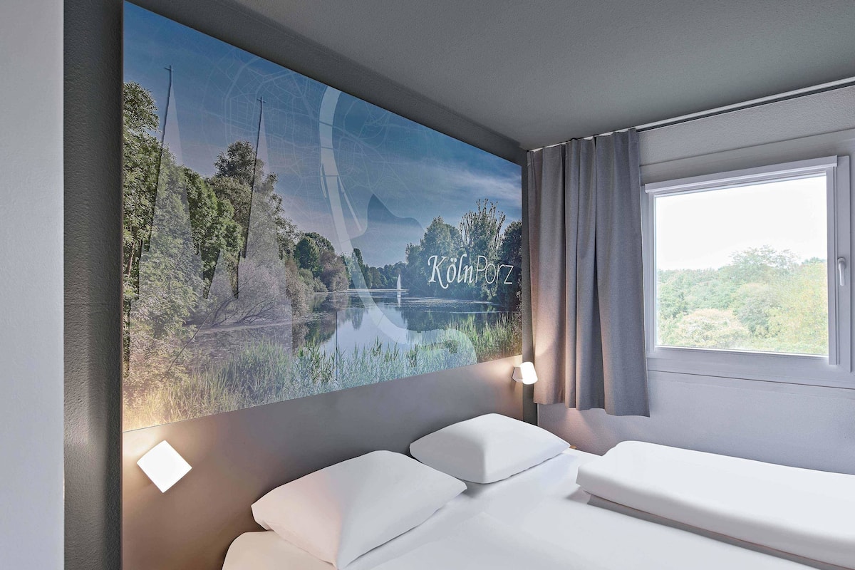 A hotel room with a bed, a nightstand, a lamp, a window with curtains, and a wall-mounted artwork of a lake and trees.