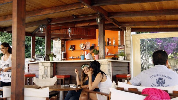 An outdoor cafe with wooden tables and chairs, a bar area with shelves of bottles, and a stone wall with a 'Welcome' sign. - Kalliopi Hotel (Minoa Pediada)