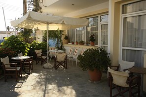 An outdoor patio with stone flooring, white and brown chairs, a table, and a large umbrella. - Kalliopi Hotel (Minoa Pediada)