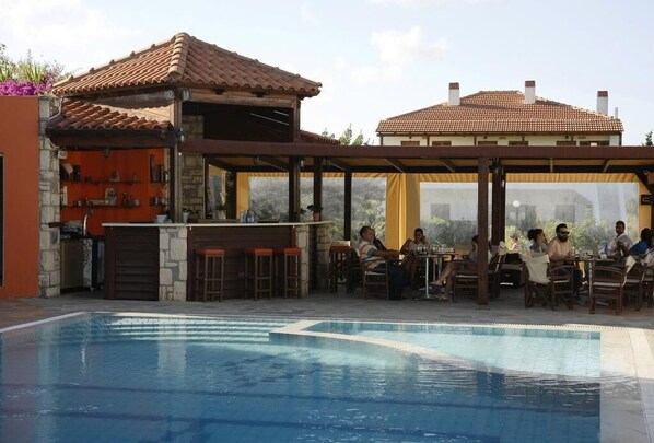 An outdoor pool area with a bar and seating, surrounded by buildings with tiled roofs. - Kalliopi Hotel (Minoa Pediada)