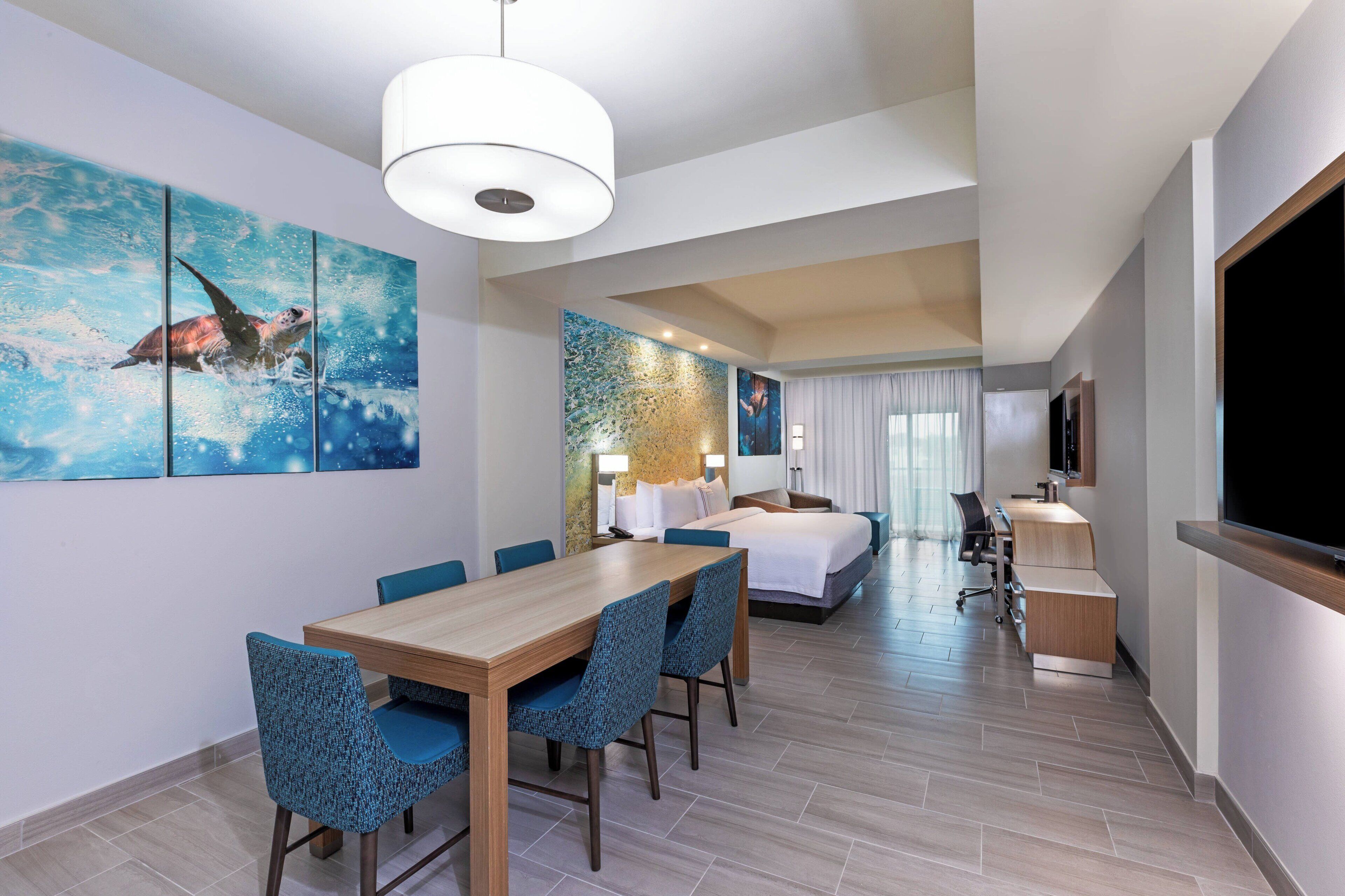Une chambre d’hôtel moderne avec une table à manger en bois, des chaises rembourrées bleues, un lit avec du linge de maison blanc, un bureau avec un ordinateur et une grande télévision.
