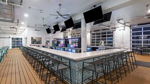 A spacious bar area with a long counter, multiple bar stools, ceiling fans, and large windows. - Courtyard by Marriott South Padre Island (South Padre Island)