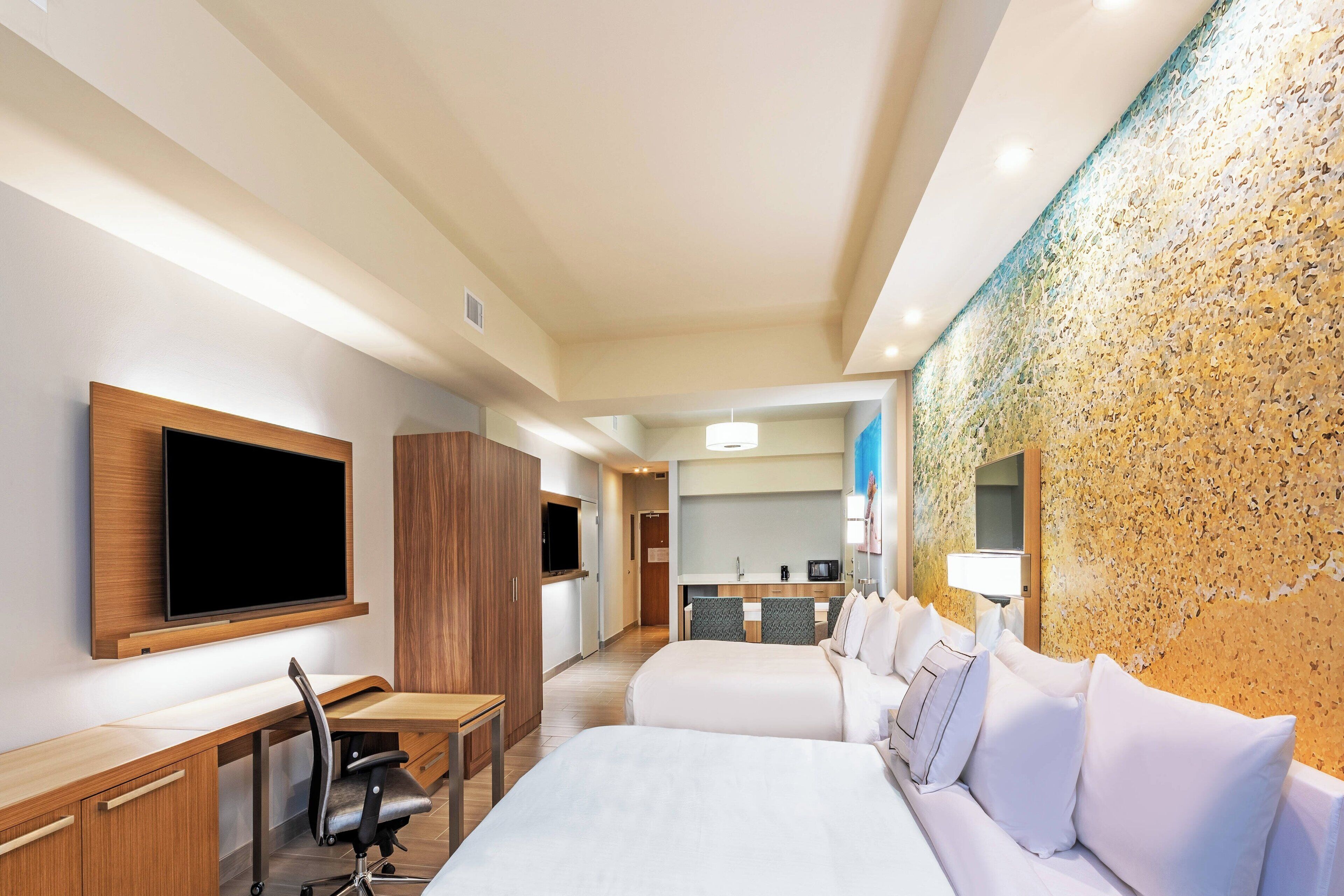 A hotel room with a bed, desk, chair, TV, and a kitchenette.