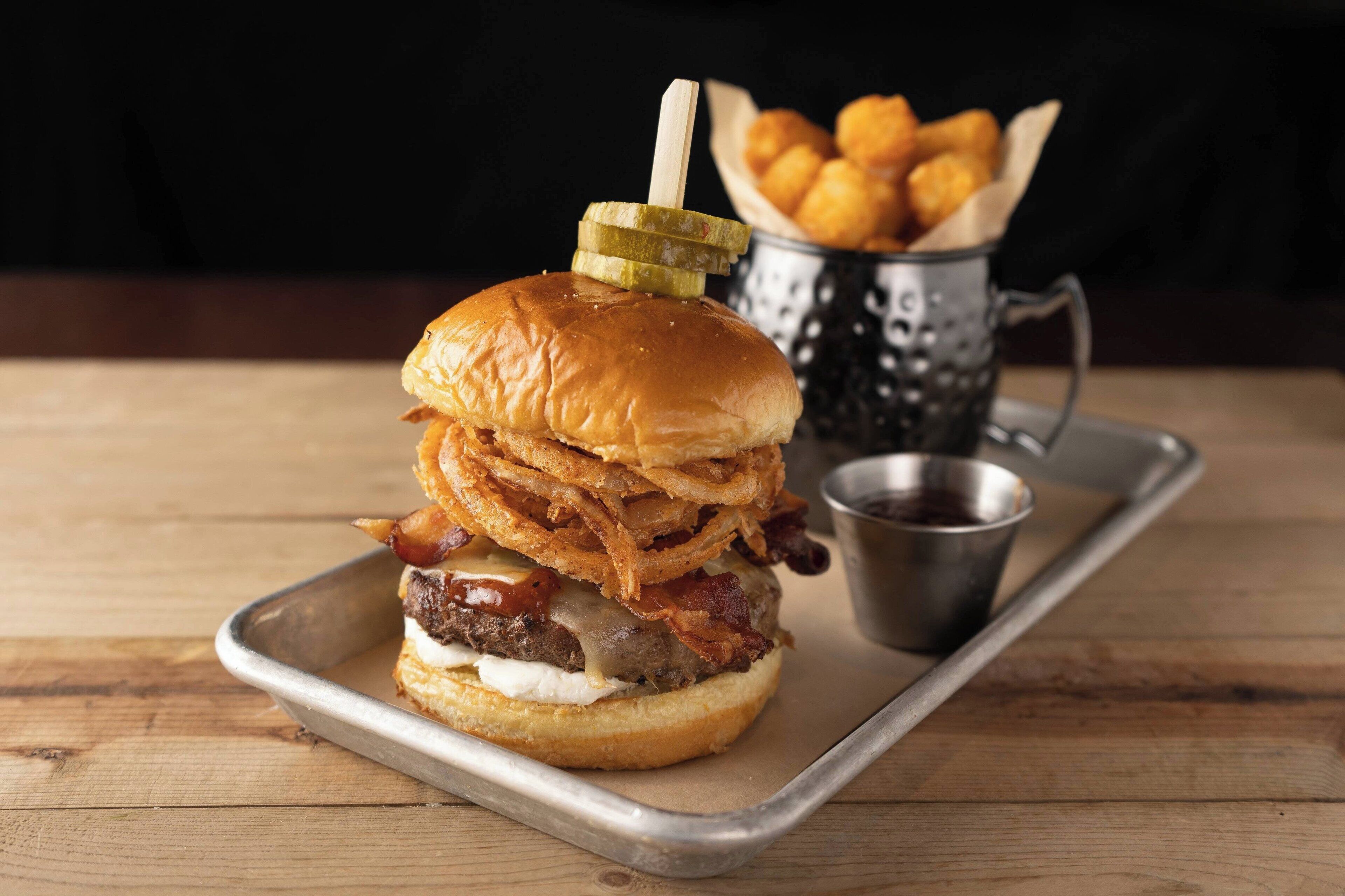 A burger with bacon, cheese, and pickles on a bun, served on a metal tray with a side of onion rings and a small cup of sauce.