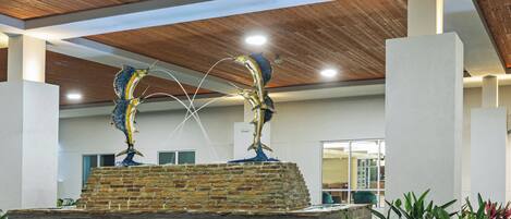 A modern indoor fountain with two fish sculptures, surrounded by plants and a brick base, under a wooden ceiling with recessed lighting.