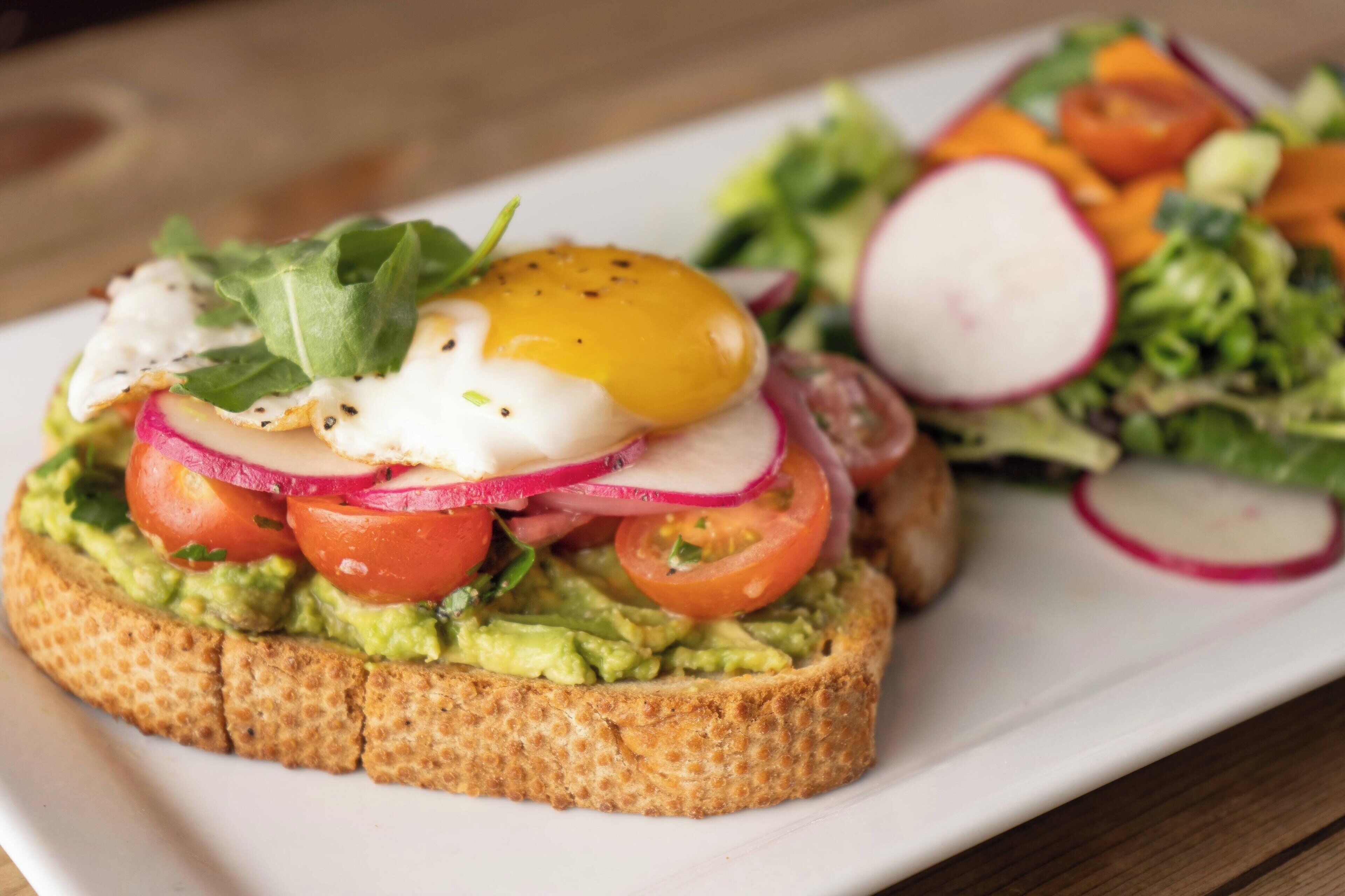 A sandwich with avocado, tomatoes, radishes, and a poached egg on a slice of bread, accompanied by a side salad.