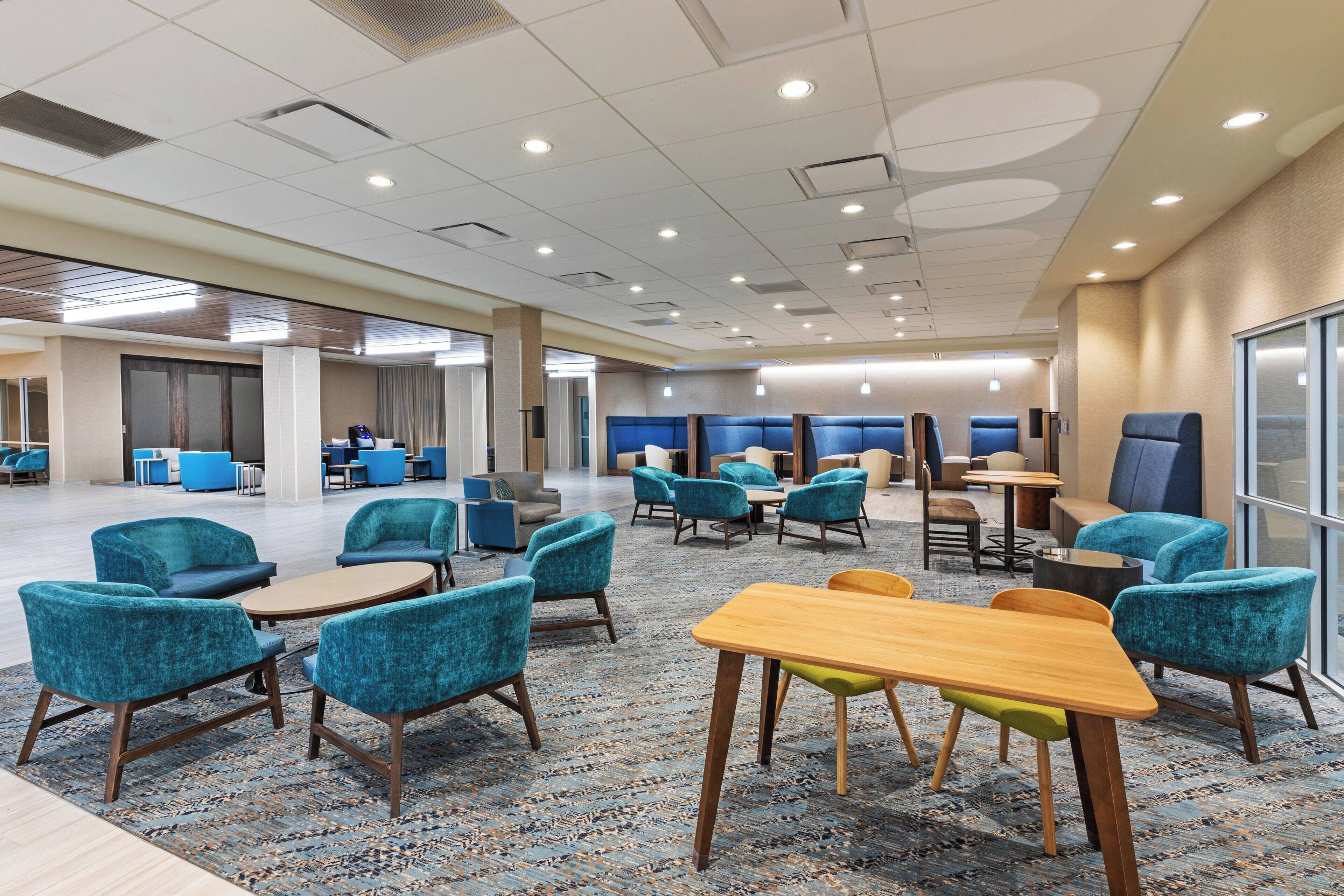 A spacious hotel lobby with multiple seating areas, including round tables and chairs, and a large window providing natural light.