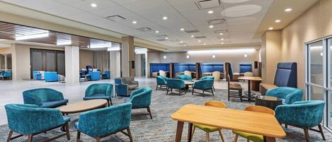A spacious hotel lobby with multiple seating areas, including round tables and chairs, and a large window providing natural light.