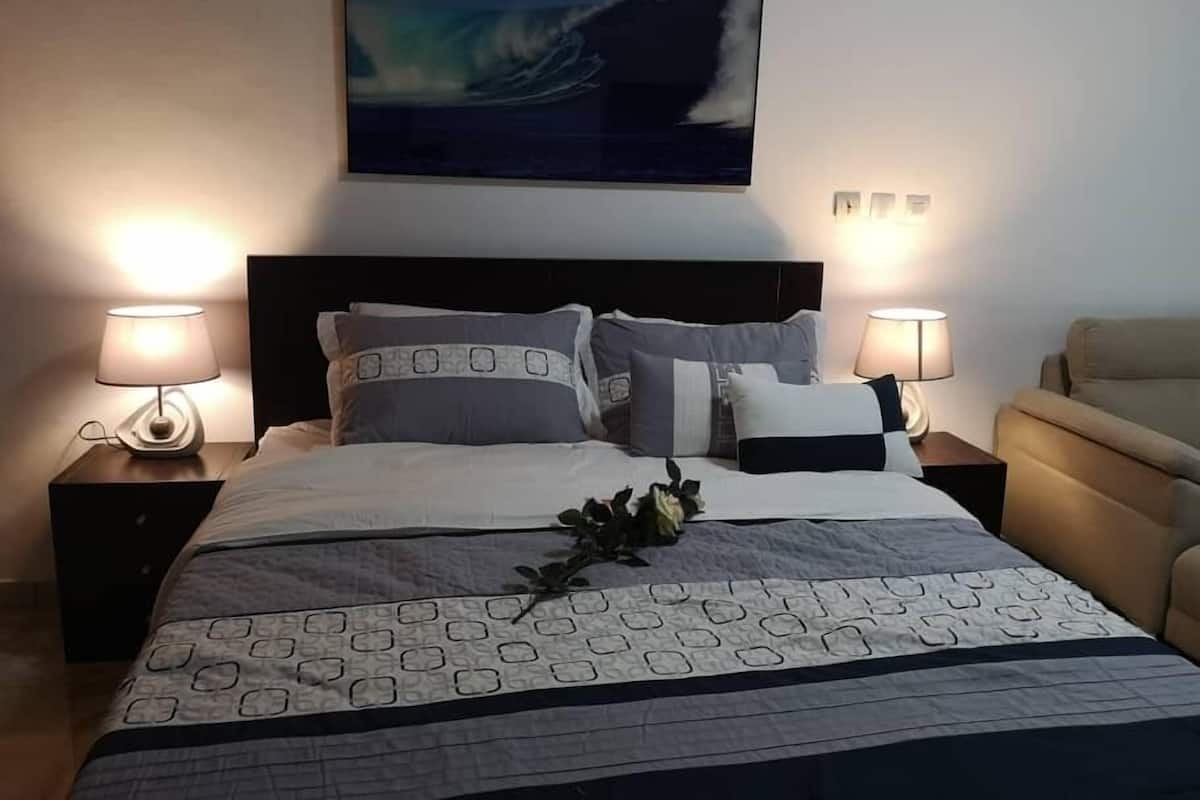 Una cama bien hecha con un edredón estampado, dos lámparas de noche y un cuadro colgado en la pared que representa una ola.